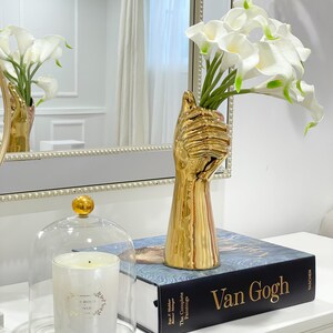 May include: A gold hand-shaped vase with white calla lilies, a glass cloche with a white candle, and a book titled "Van Gogh: The Complete Paintings" by Taschen.
