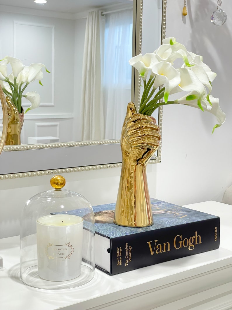 May include: A gold hand-shaped vase with white calla lilies, a glass cloche with a white candle, and a book titled "Van Gogh: The Complete Paintings" by Taschen.
