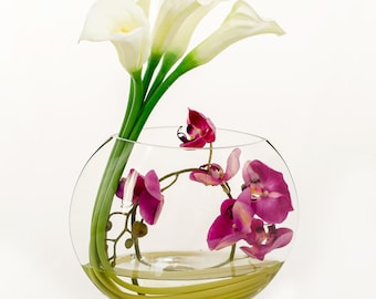 Uniek ontwerp, grote, realistische, elegante witte calla lelies en paarse orchideeën in een halfronde glazen vaas. Kunstmatige imitatie arrangement.