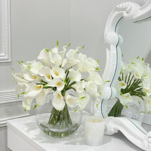 May include: A white vase filled with a bouquet of white calla lilies. The vase is sitting on a white table with a white candle in front of it.