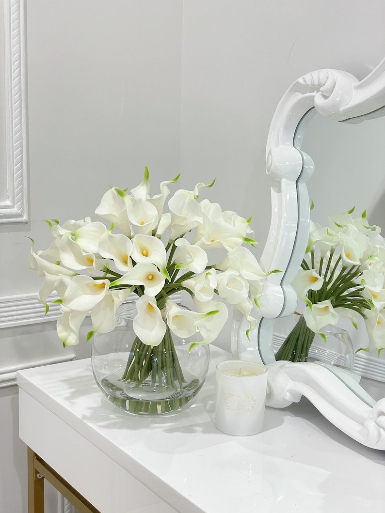 May include: A white vase filled with a bouquet of white calla lilies. The vase is sitting on a white table with a white candle in front of it.