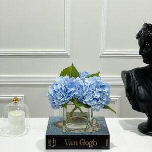 May include: A blue hydrangea flower arrangement in a clear glass vase on top of a book titled "Van Gogh" with the text "The Complete Paintings" on the spine.