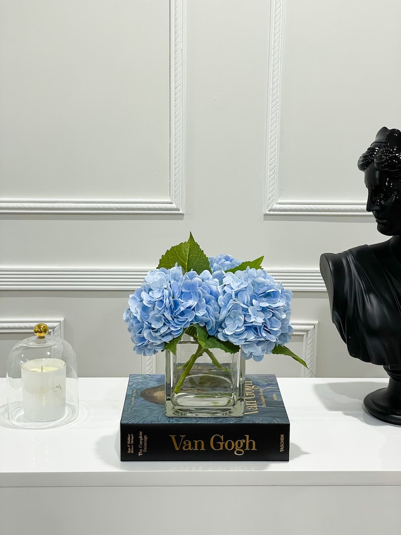May include: A blue hydrangea flower arrangement in a clear glass vase on top of a book titled "Van Gogh" with the text "The Complete Paintings" on the spine.