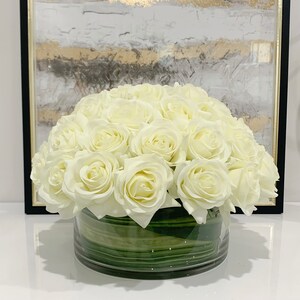 May include: A bouquet of white artificial roses arranged in a clear glass vase with green leaves. The vase has a textured, striped pattern.