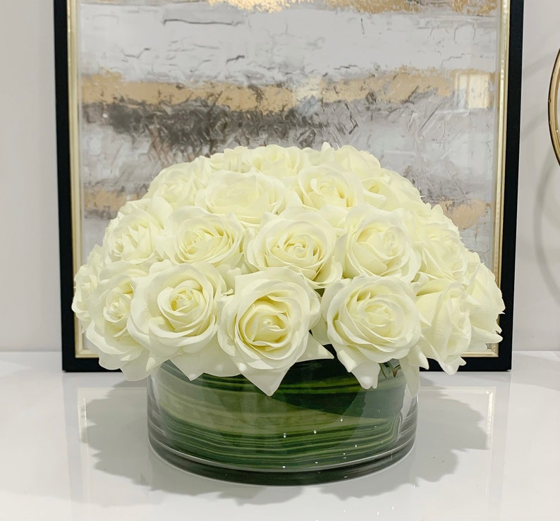 May include: A bouquet of white artificial roses arranged in a clear glass vase with green leaves. The vase has a textured, striped pattern.