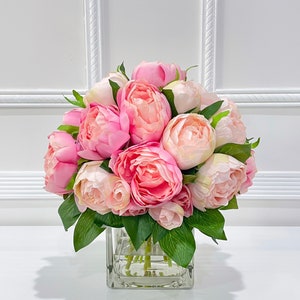K&ouml;nnte beinhalten: Ein Strau&szlig; rosa Pfingstrosen, die in einer klaren Glasvase arrangiert sind. Die Pfingstrosen sind in voller Bl&uuml;te und haben ein weiches, zartes Aussehen.