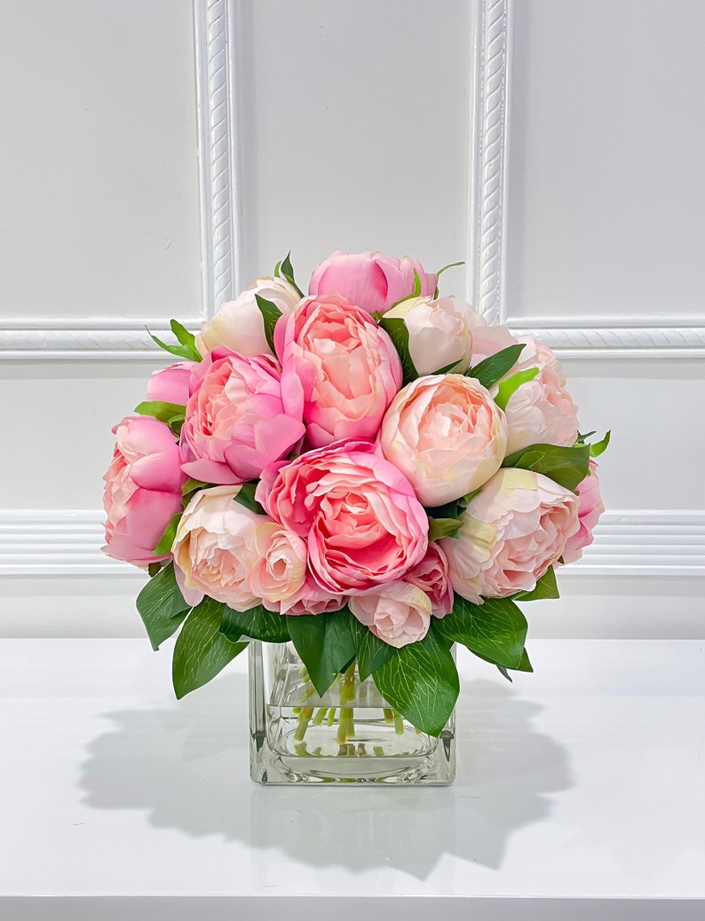 K&ouml;nnte beinhalten: Ein Strau&szlig; rosa Pfingstrosen, die in einer klaren Glasvase arrangiert sind. Die Pfingstrosen sind in voller Bl&uuml;te und haben ein weiches, zartes Aussehen.