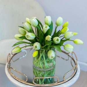 May include: A white marble side table with a gold metal base and a mirrored top. A clear glass vase with a bouquet of white and green tulips sits on the table.