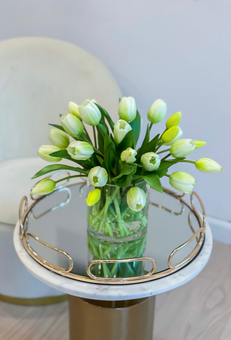 May include: A white marble side table with a gold metal base and a mirrored top. A clear glass vase with a bouquet of white and green tulips sits on the table.