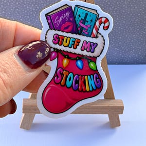 May include: A die-cut sticker shaped like a pink Christmas stocking with a white fur cuff. The stocking features the text "STUFF MY STOCKING" in colorful letters, a string of multi-colored lights, two books, and a red and white candy cane.
