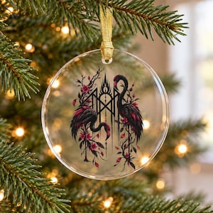 May include: A circular clear acrylic ornament featuring a printed illustration of two black flamingos with pink accents, surrounded by thorny branches and small pink flowers, suspended by a gold ribbon loop for hanging.