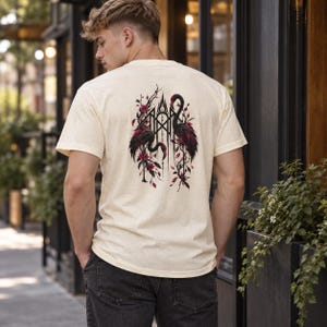 May include: A cream-colored short-sleeve t-shirt featuring a graphic on the back. The design displays two dark-colored flamingos facing each other, surrounded by intricate black geometric lines and deep red floral accents and foliage.