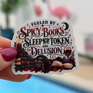 May include: A die-cut sticker featuring the text "FUELED BY SPICY BOOKS SLEEP TOKEN AND DELUSION" in black and red script. The design includes a stack of books, a lit candle, headphones, an open book, a wine glass, and red roses.