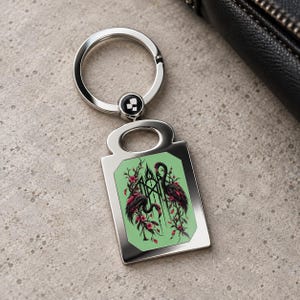 May include: A silver-toned metal keychain featuring a rectangular pendant with a light green background. The pendant displays a graphic illustration of two dark flamingos surrounded by pink flowers and black geometric line art.