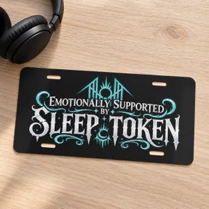 May include: A rectangular black license plate featuring the white and teal text "EMOTIONALLY SUPPORTED BY SLEEP TOKEN" in a stylized font. The design includes intricate decorative flourishes, star accents, and a central crescent moon symbol.