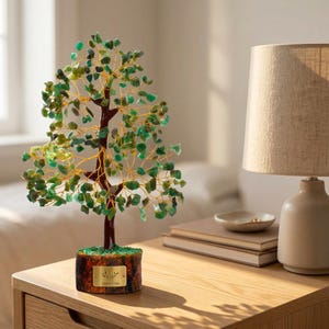 May include: A decorative tree sculpture featuring a dark brown trunk and thin gold wire branches adorned with numerous small, translucent green stone chips. The tree is mounted on a cylindrical wooden base with a small gold rectangular plaque.