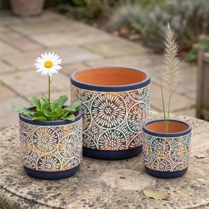 May include: Three cylindrical ceramic planters in varying sizes featuring a dark blue rim and base with a repeating white and gold circular floral pattern. The interior of each planter is a natural terracotta orange color.