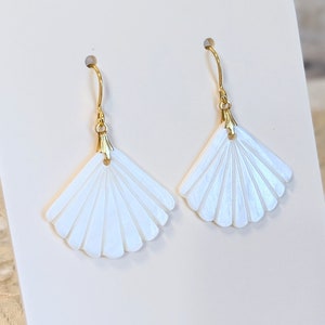 May include: A pair of white, fan-shaped earrings with gold-coloured hooks. The earrings are displayed on a white stand against a blurred background. The earrings have a pearlescent sheen and are intricately detailed.