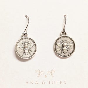 May include: A pair of silver-toned bee earrings. Each earring features a circular pendant with a detailed bee design. The earrings are displayed on a white card with the text "ANA & JULES" at the bottom.