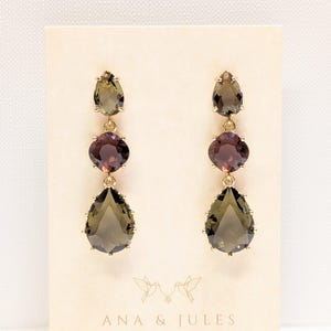 May include: A pair of drop earrings featuring three faceted stones in gold-tone settings. Each earring has a small pear-shaped olive stone at the top, a round deep red stone in the center, and a large pear-shaped olive stone at the base.