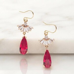 May include: A pair of gold-toned drop earrings featuring a fan-shaped top section adorned with clear baguette-cut stones and a single round stone, suspending a vibrant, pear-shaped red gemstone. The earrings hang from simple gold-toned wire hooks.