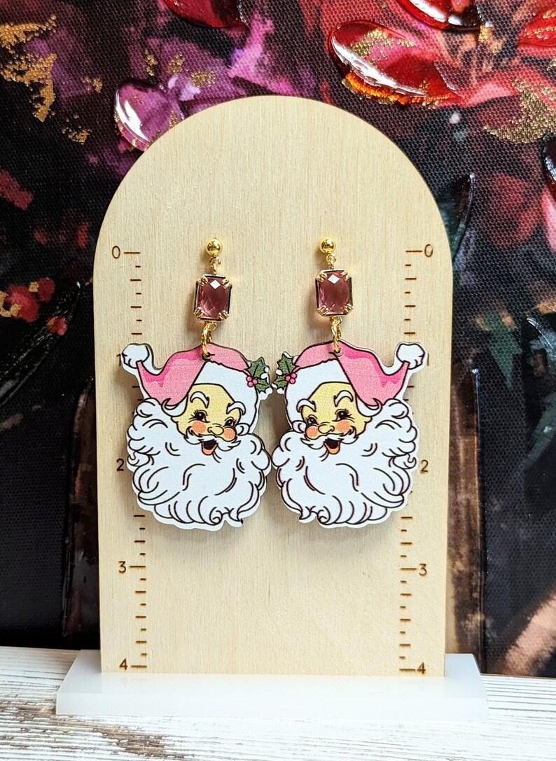 May include: A pair of festive earrings with a Santa Claus design. Each earring has a gold-coloured top with a rectangular pink gemstone, and a Santa Claus face with a pink hat and white beard. The earrings are displayed on a wooden stand.