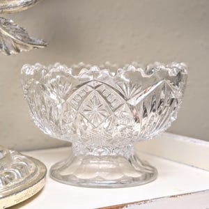 May include: A clear, cut-glass bowl with a pedestal base. The bowl features a detailed, geometric pattern and a scalloped rim. The glass reflects light, highlighting the intricate design. This decorative bowl is suitable for serving or display.