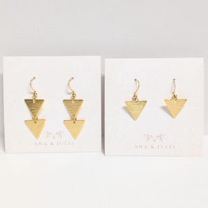 May include: Two pairs of gold-colored earrings. One pair features two stacked triangles, and the other pair has a single triangle. Both pairs are on white cards with the text "ANA & JULES".