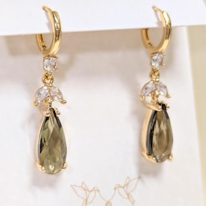 May include: Gold-coloured earrings with olive-green, teardrop-shaped gemstones. Each earring has a cluster of clear crystals and a hinged hoop. The design is elegant and suitable for various occasions.