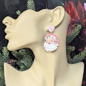 May include: Pink and white Santa Claus earrings. The earrings feature a heart-shaped stud with a pink and white checkered pattern. The Santa Claus pendant has a pink hat and a white beard. The earrings are a festive accessory.