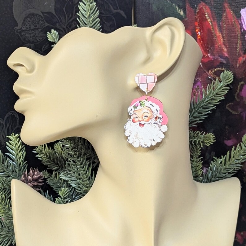 May include: Pink and white Santa Claus earrings. The earrings feature a heart-shaped stud with a pink and white checkered pattern. The Santa Claus pendant has a pink hat and a white beard. The earrings are a festive accessory.