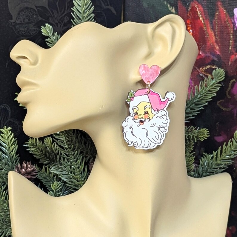 May include: Pink Santa Claus earrings with a heart-shaped top. The earrings feature a cartoon Santa Claus with a pink hat and a white beard. The earrings are attached to a mannequin head.