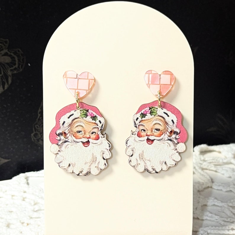 May include: A pair of Christmas-themed earrings featuring Santa Claus faces. The earrings have pink heart-shaped tops with a grid pattern, and the Santa faces have white beards and pink hats decorated with holly.