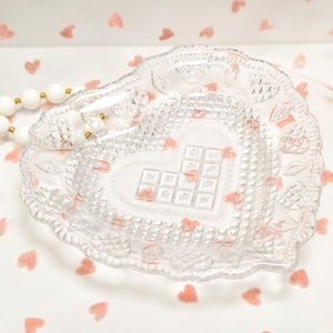 May include: A clear, heart-shaped glass dish with a textured surface and a decorative border. The dish has a heart-shaped design in the center with pink accents. A white beaded necklace with gold accents is visible.