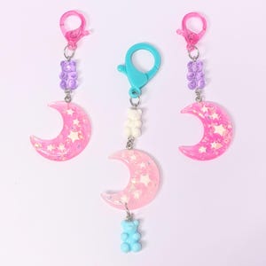 May include: Three keychains with pink crescent moon charms, star accents, and gummy bear beads. The charms have pink and white glitter. Each keychain has a different color clasp and gummy bear.