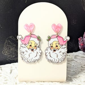 May include: A pair of Santa Claus earrings. Each earring has a pink heart-shaped top and a Santa Claus face with a pink hat, white beard, and a smiling expression. The earrings are designed with a vintage aesthetic.