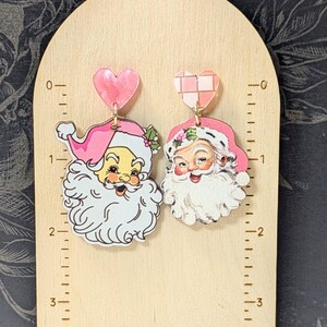 May include: A pair of Christmas-themed earrings featuring Santa Claus. Each earring has a vintage Santa face with a pink hat and white beard. They hang from pink heart-shaped studs.