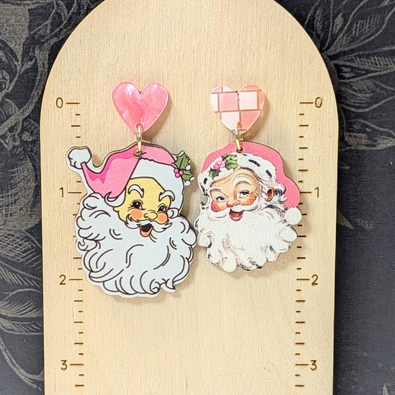 May include: A pair of Christmas-themed earrings featuring Santa Claus. Each earring has a vintage Santa face with a pink hat and white beard. They hang from pink heart-shaped studs.