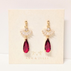 May include: Gold-toned drop earrings with a fan-shaped top adorned with clear crystals and a teardrop-shaped, deep red gemstone. The earrings are displayed on a cream-colored card with the text "ANA & JULES" and a bird graphic.