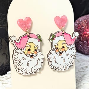 May include: A pair of festive earrings featuring Santa Claus faces with pink hats and heart-shaped tops. The Santa faces have white beards and rosy cheeks. The earrings are designed with gold-coloured hooks.
