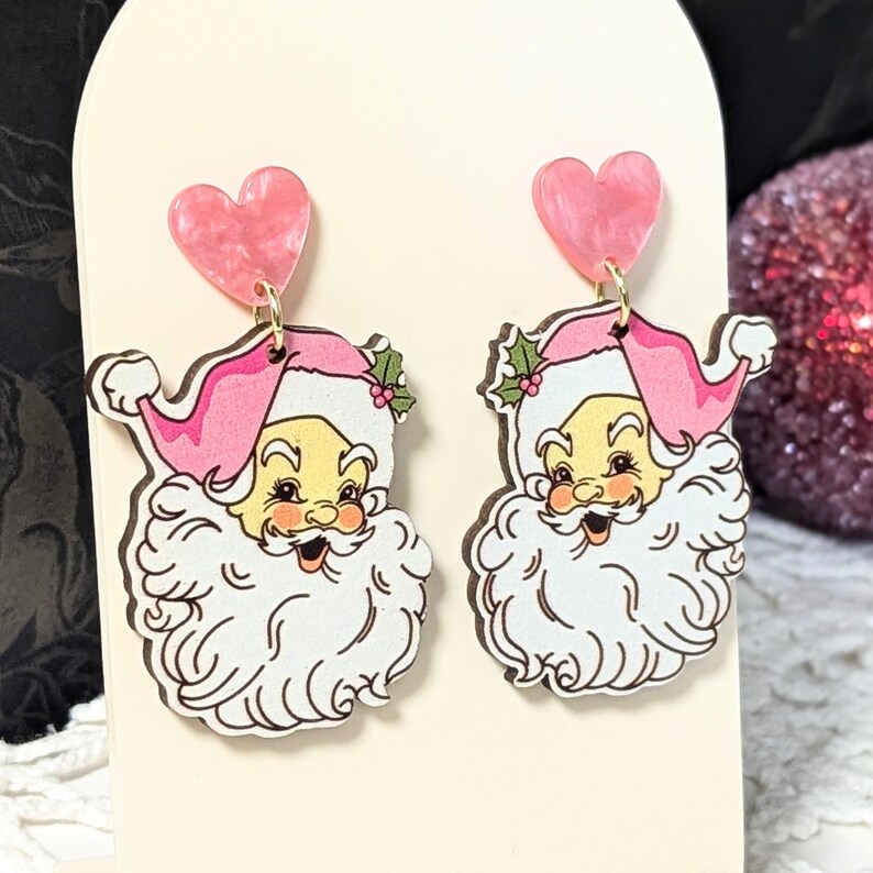 May include: A pair of festive earrings featuring Santa Claus faces with pink hats and heart-shaped tops. The Santa faces have white beards and rosy cheeks. The earrings are designed with gold-coloured hooks.
