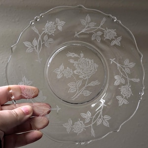 May include: Clear glass plate with a scalloped edge, featuring an etched rose and leaf design. The plate has a central rose motif and floral patterns around the rim. The plate is held by a hand.