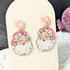 May include: A pair of Santa Claus earrings. Each earring features a smiling Santa face with a white beard and pink hat. The earrings are connected to a pink and white checkered heart-shaped stud with a gold-coloured hook.