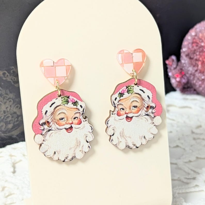 May include: A pair of Santa Claus earrings. Each earring features a smiling Santa face with a white beard and pink hat. The earrings are connected to a pink and white checkered heart-shaped stud with a gold-coloured hook.