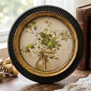 May include: A circular wall frame featuring a black fluted outer edge and a gold inner rim. The artwork inside depicts a bouquet of white flowers in a glass vase against a cream-colored background, capturing a still life botanical illustration.