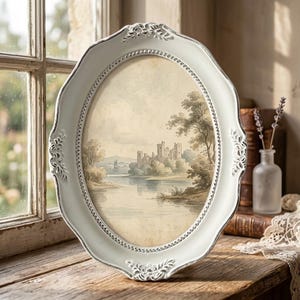May include: An oval picture frame with a distressed white finish and ornate, raised floral detailing along the edges. The frame contains a landscape print depicting a stone castle situated on the bank of a calm river surrounded by trees.