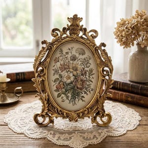 May include: An oval picture frame with an ornate gold-colored finish featuring intricate scrollwork and leaf motifs. The frame contains a botanical print of a colorful wildflower bouquet on a cream background, supported by a decorative base.