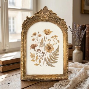 May include: An arched gold picture frame with ornate detailing containing a botanical print. The print features a collection of pressed-style flowers, leaves, and a fern frond in shades of sepia, tan, and brown on a cream-coloured background.