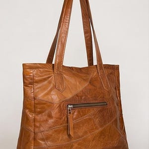 Peut inclure: Un sac cabas en cuir marron clair présentant un motif en patchwork avec des coutures apparentes. Le sac comprend deux longues anses et une poche extérieure zippée horizontale avec une tirette en cuir, affichant une finition texturée.