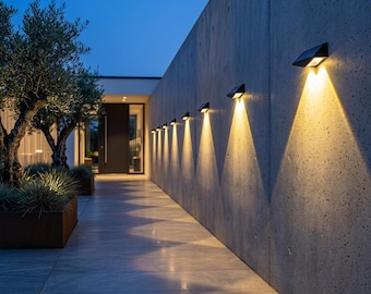 May include: A series of black, angular wall-mounted outdoor light fixtures attached to a smooth concrete wall. Each fixture emits a bright, warm, triangular beam of light directed downward, illuminating the gray tiled walkway below at dusk.
